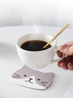 A grey-and-white cat coaster under a steaming coffee cup on a white table