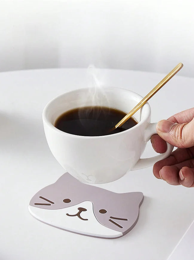 A grey-and-white cat coaster under a steaming coffee cup on a white table