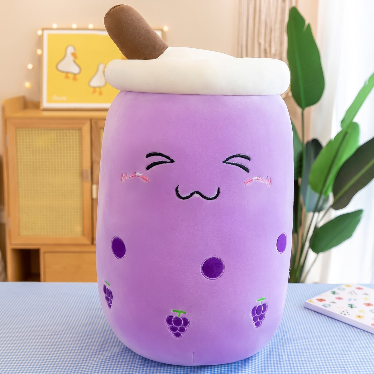 Boba Plush Pillow in purple with grape prints, closed happy eyes and blushing cheeks, a plush bubble-tea cup with a cream lid, straw and pearls