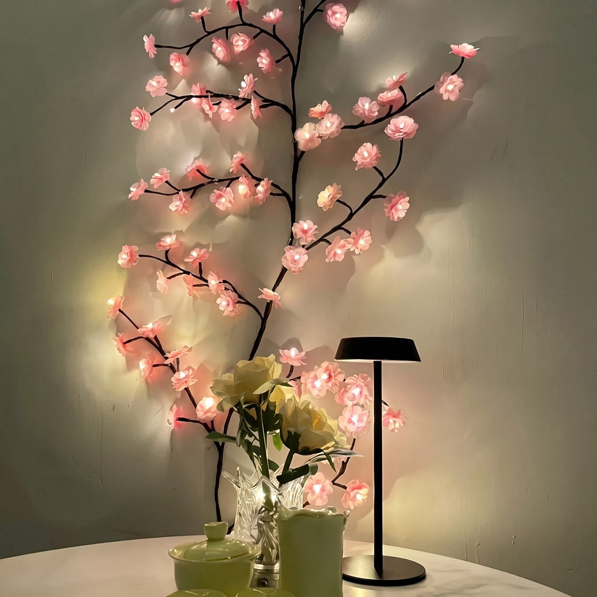 Pink Cherry Blossom Branch Light curving up a wall beside a table lamp and flowers