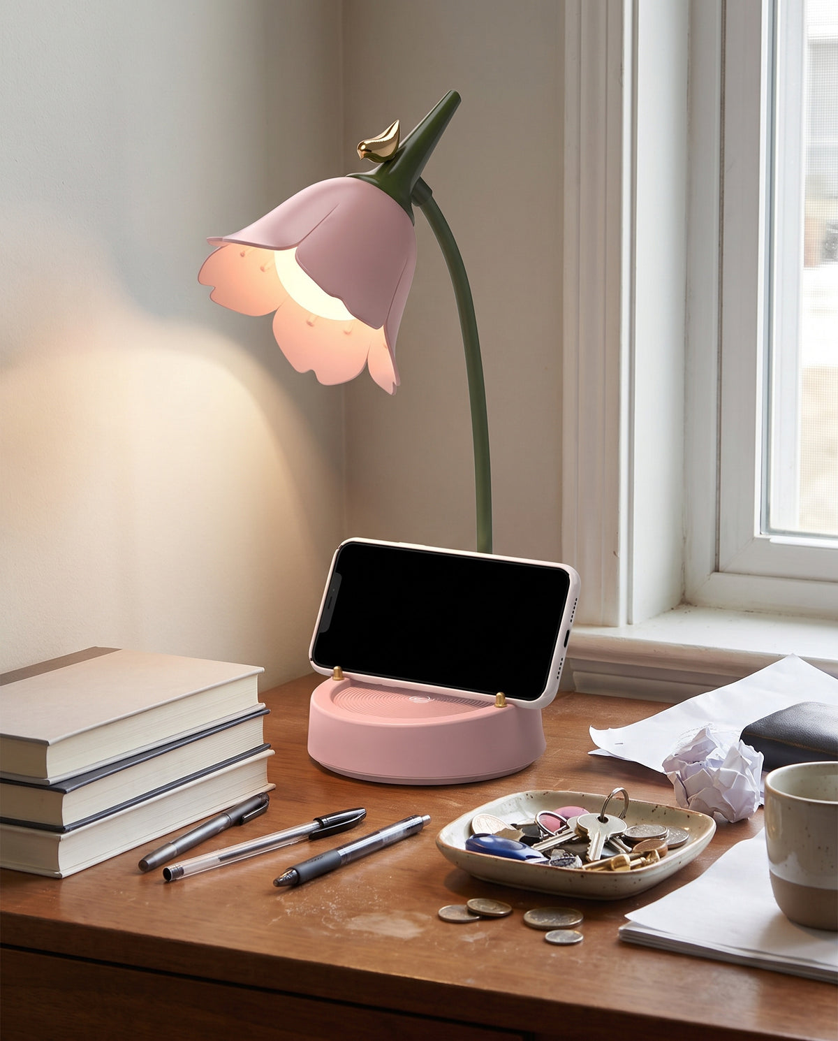 Flower LED Desk Lamp in pink, lit warm on a wooden desk, with a phone standing in the base's holder groove