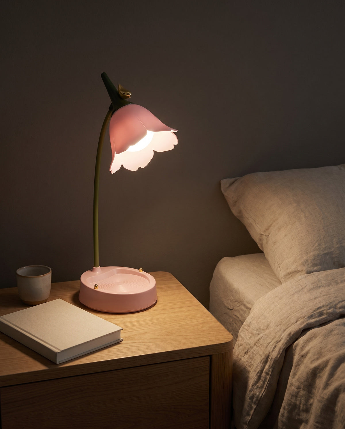 Flower LED Desk Lamp in pink, lit warm on a nightstand at night, beside a bed