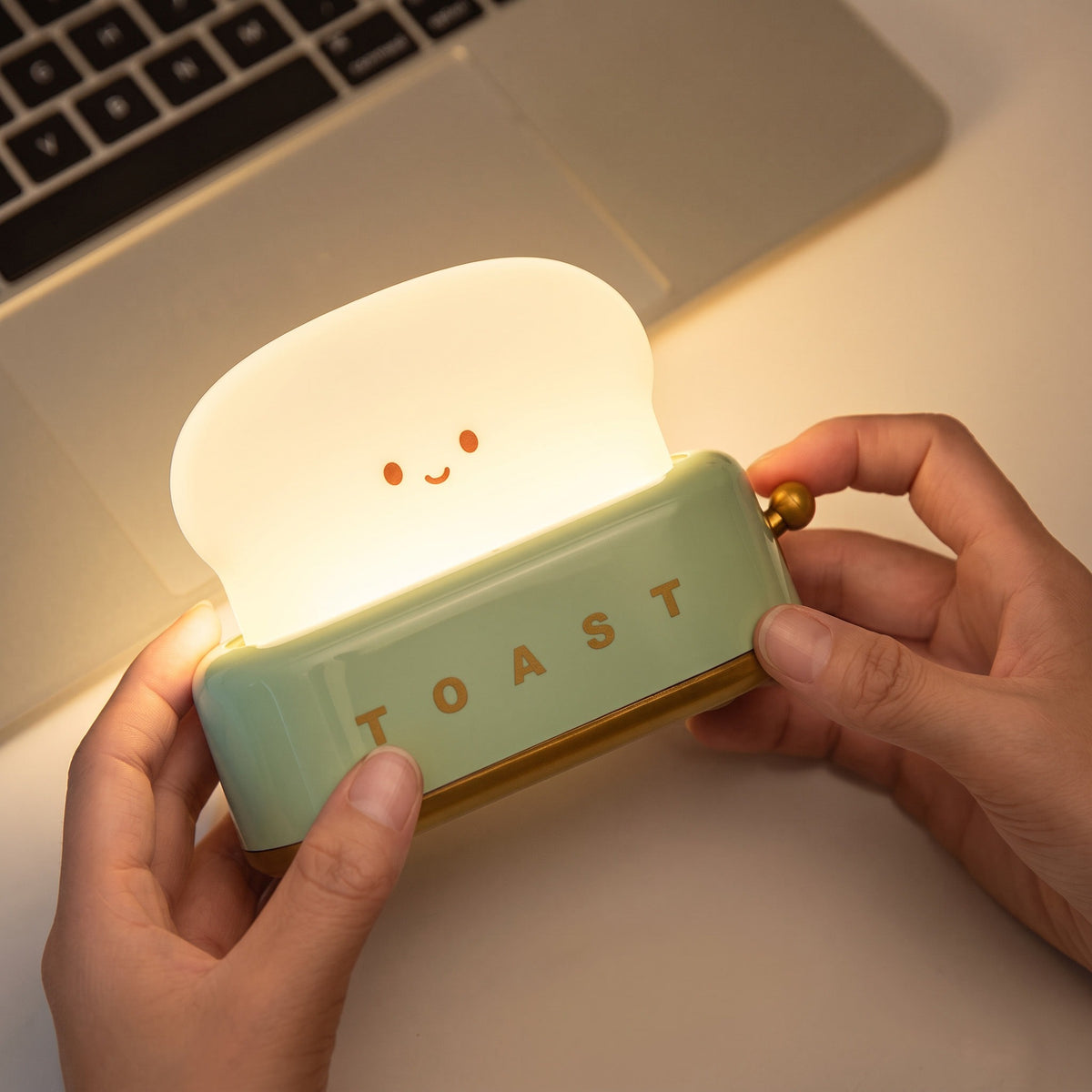 Toaster Night Light in mint, lit, held in two hands beside a laptop to show its palm size